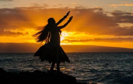 Silhouetted young woman in traditional costume, hula dancing on coastal rock at sunset, Maui, Hawaii, USA