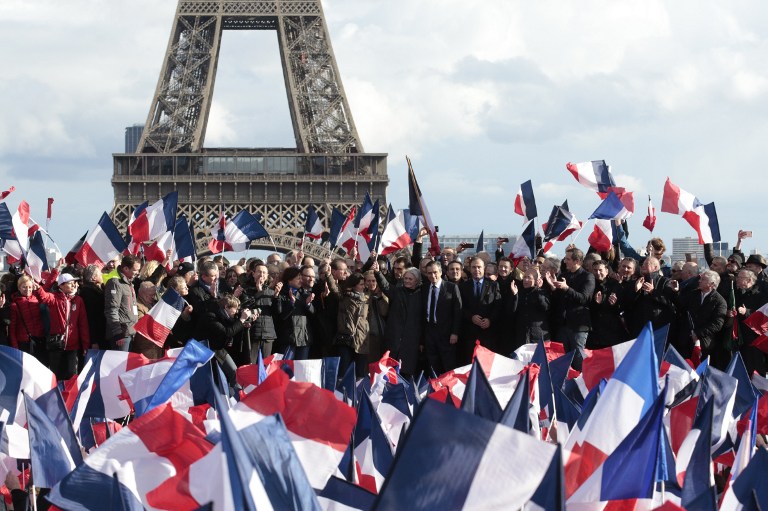 French presidential election candidate for the right-wing Les Republicains (LR) party Francois Fillon (C), flanked by his wife Penelope Fillon and his daughter Marie Fillon (his L side), acknowledges the audience as he stands on stage with the Eiffel tower on the background, while his supporters greet him with French national flags during a rally at the place du Trocadero, in Paris, on March 5, 2017. Embattled French conservative Francois Fillon told supporters at a Paris rally on March 5, 2017 to "never give up the fight" as he strives to stay in the presidential election race amid an expenses scandal. Fillon, who is to be charged over claims he gave his wife and children highly-paid fake parliamentary jobs, told the rain-drenched crowd he had been "attacked by everyone" in the campaign. / AFP PHOTO / GEOFFROY VAN DER HASSELT